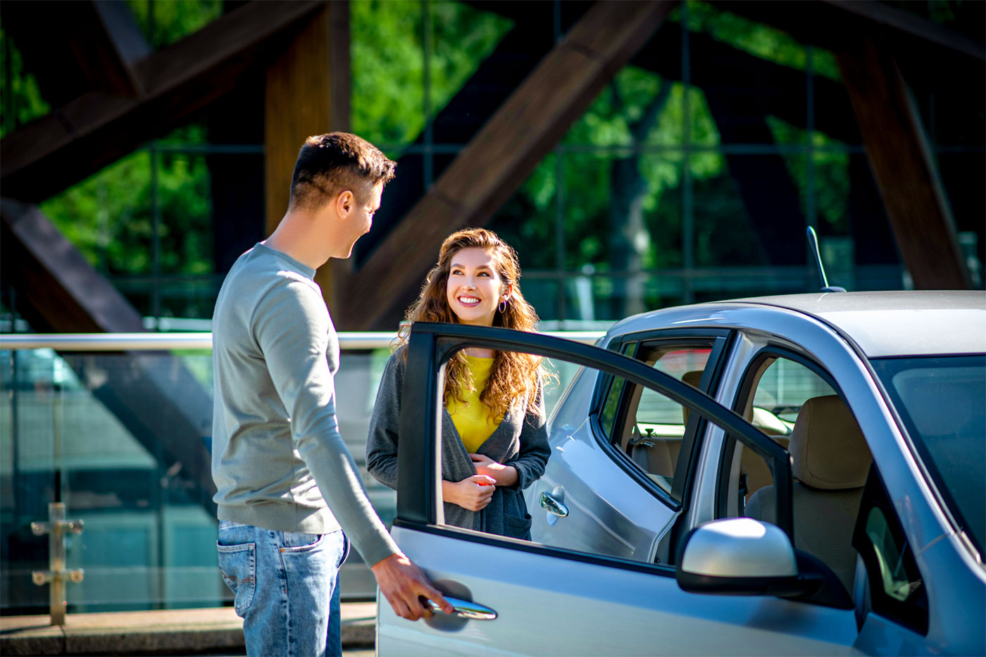 Ein Mann &ouml;ffnet einer Frau die Autot&uuml;r auf einem Parkplatz, beide stehen neben einem silberfarbenen Auto.