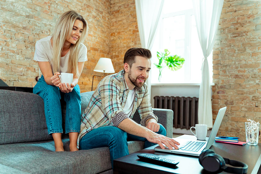 Ein Mann sitzt mit einem Laptop auf dem Sofa, eine Frau mit einer Tasse steht daneben und beide schauen auf den Bildschirm eines Laptops.