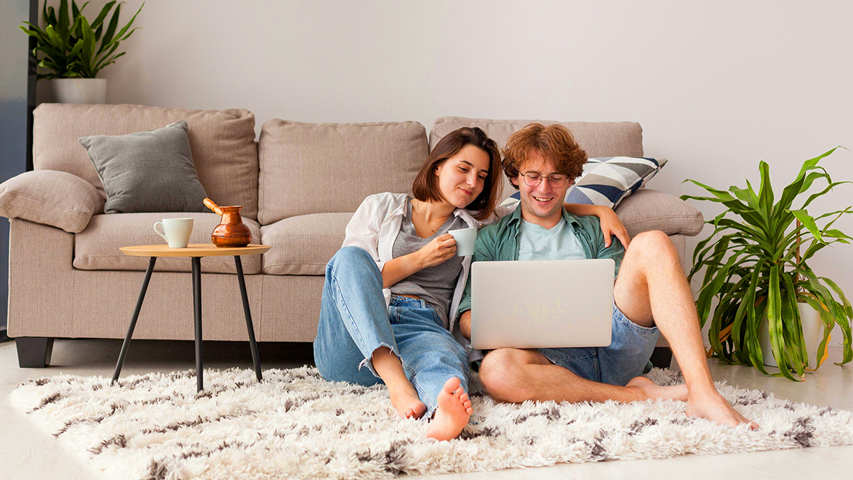  Zwei Personen sitzen entspannt auf einem Teppich vor einem Sofa und schauen gemeinsam auf einen Laptop. Eine Person hat den Arm um die andere gelegt und h&auml;lt eine Tasse. Neben ihnen stehen Zimmerpflanzen und ein kleiner Beistelltisch mit Geschirr. Die Szene vermittelt eine ruhige, gem&uuml;tliche Atmosph&auml;re und gemeinsames Arbeiten oder Anschauen von Inhalten am Computer.