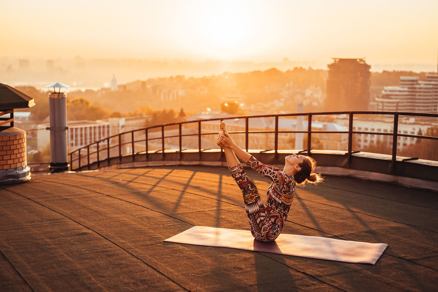 Frau auf Yogamatte macht Yogaübung auf einem Dach mit Stadt im Hintergrund bei Sonnenaufgang oder Sonnenuntergang.