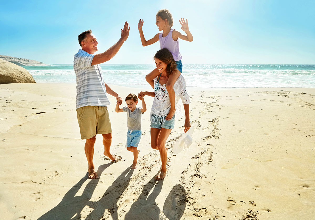 Eine Familie l&auml;uft gemeinsam barfu&szlig; am Strand entlang. Ein Kind sitzt auf den Schultern einer Frau. Ein Mann gibt dem Kind ein High Five. Ein weiteres kleines Kind wird an der Hand gef&uuml;hrt. Im Hintergrund sind das Meer und sanfte Wellen zu sehen.