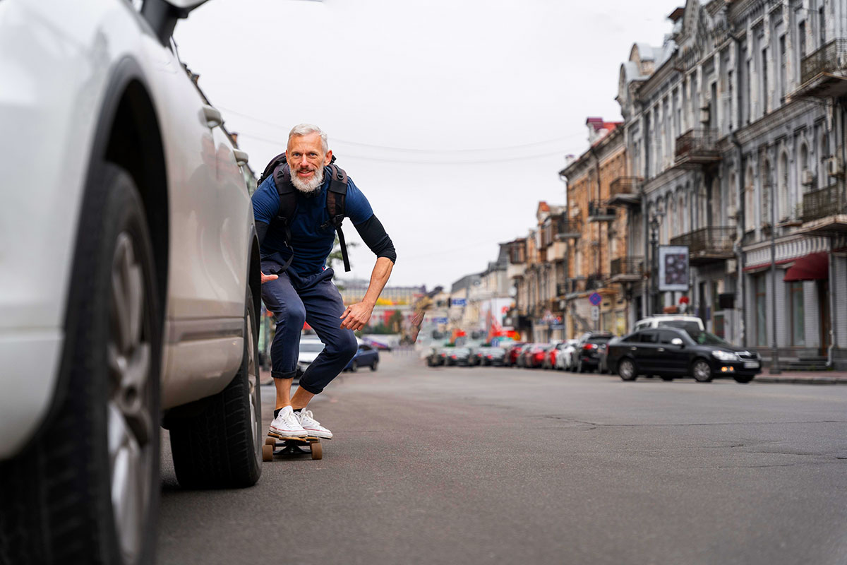 Eine Person f&auml;hrt auf einem Skateboard durch eine st&auml;dtische Stra&szlig;e zwischen parkenden Autos. Die Person tr&auml;gt einen Rucksack und bewegt sich in gebeugter Haltung auf der Fahrbahn. Im Hintergrund stehen mehrst&ouml;ckige Geb&auml;ude entlang der Stra&szlig;e.