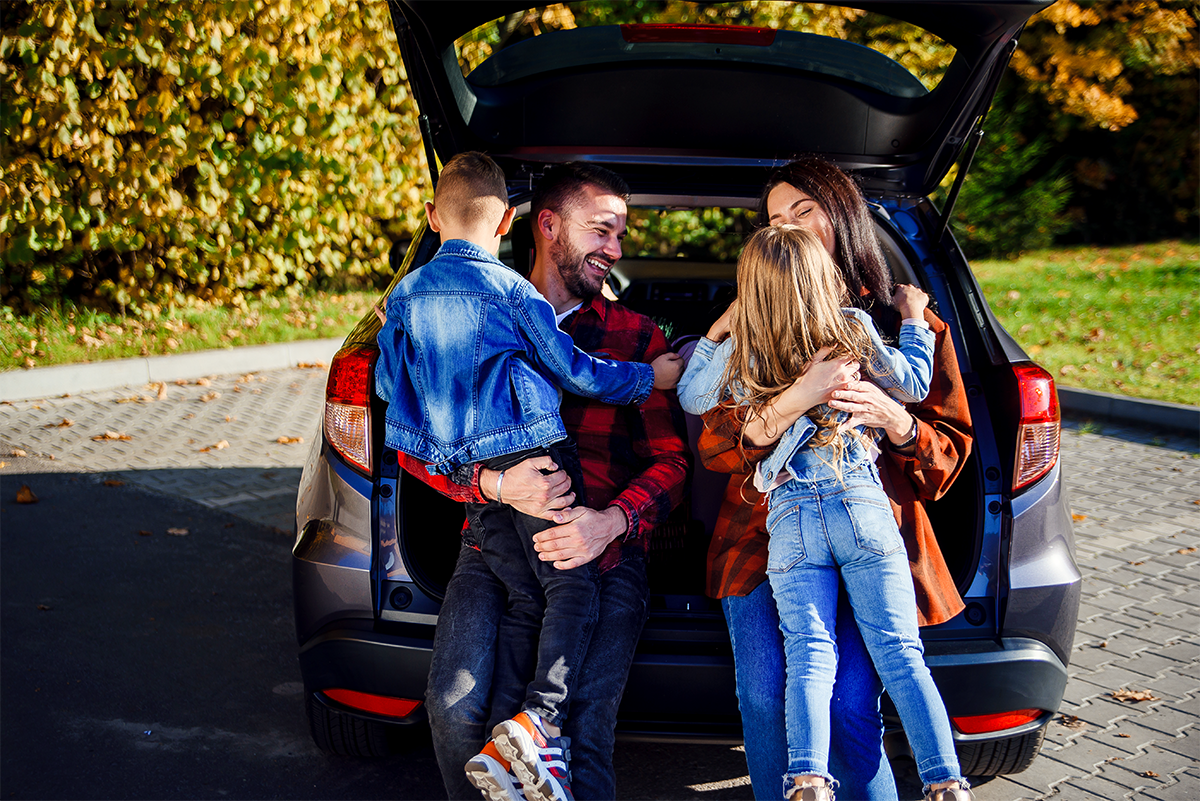 Vierk&ouml;pfige Familie sitzt auf der ge&ouml;ffneten Kofferraumklappe eines Autos, zwei Erwachsene und zwei Kinder umarmen sich.