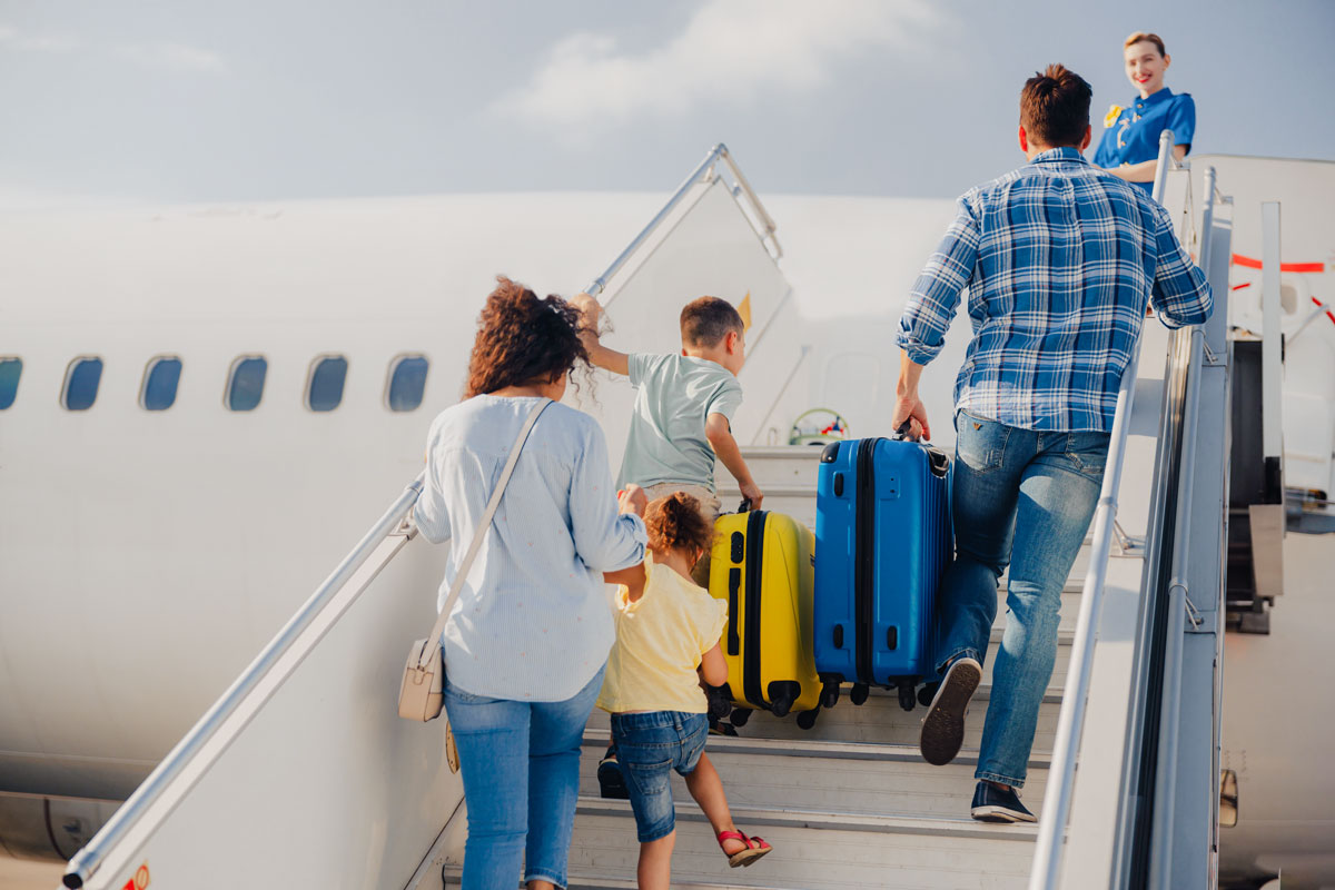 Eine Familie mit zwei Kindern steigt eine Flugzeugtreppe hinauf, der Vater trägt einen Koffer, der Junge trägt ebenfalls einen Koffer, die Flugbegleiterin steht oben am Eingang des Flugzeugs