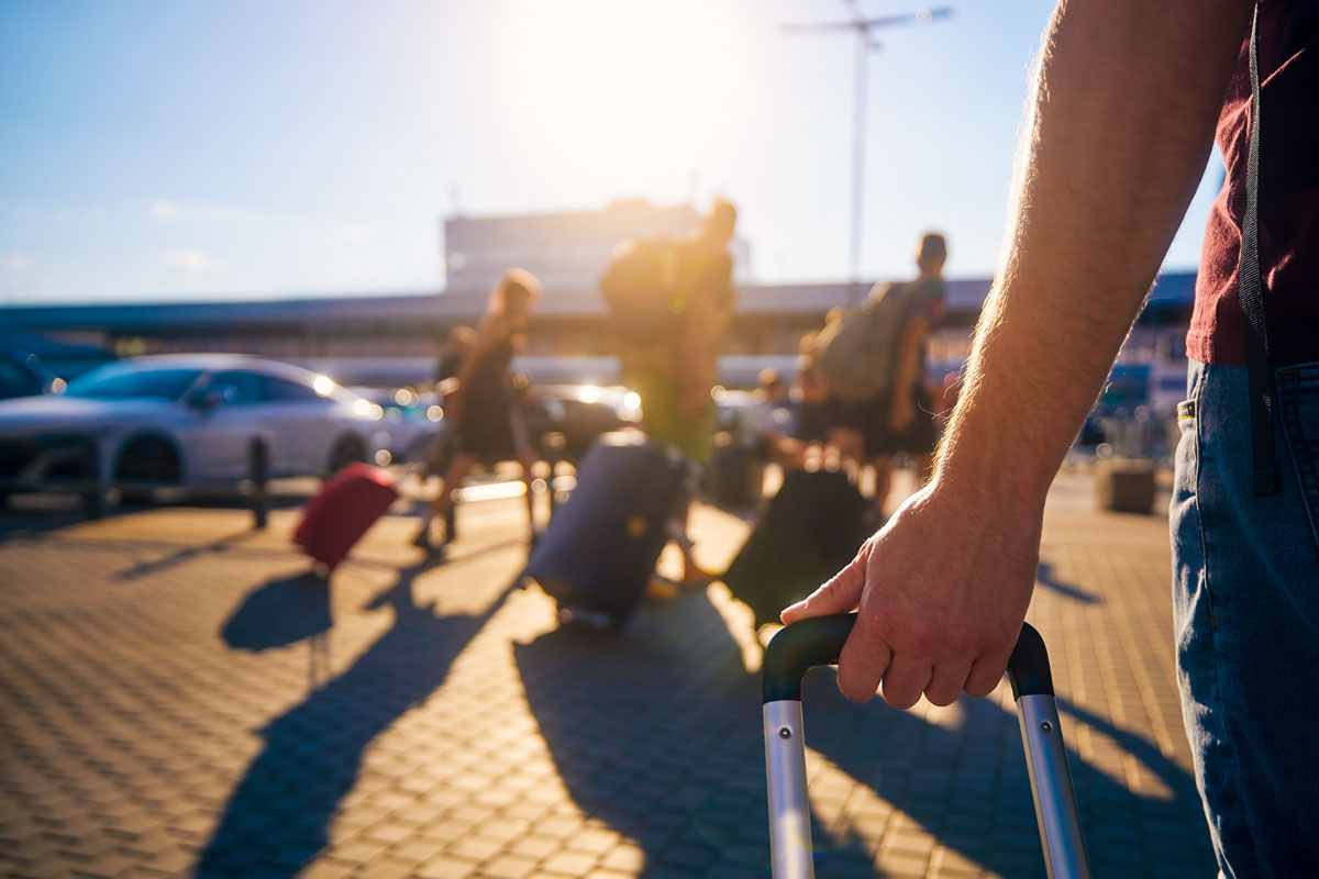 Nahaufnahme einer Hand, die den Griff eines Rollkoffers hält, im Hintergrund unscharfe Personen mit Gepäck auf einem gepflasterten Parkplatz mit geparkten Autos bei Sonnenschein.