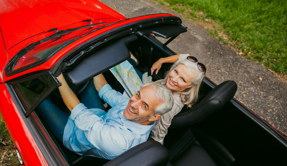 ipb_blog_hitze_1 Ein älteres Paar sitzt in einem Cabrio. Sie schauen lächelnd nach oben in die Kamera. Auf dem Schoß der Frau, die auf dem Beifahrersitz sitzt, liegt eine Landkarte.