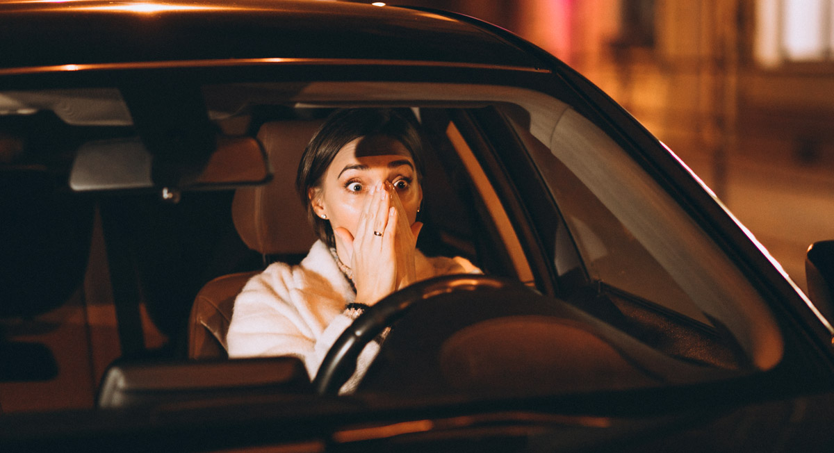 Frau mit erschrockenem Gesichtsausdruck sitzt am Steuer eines Autos und hält eine Hand vor den Mund.