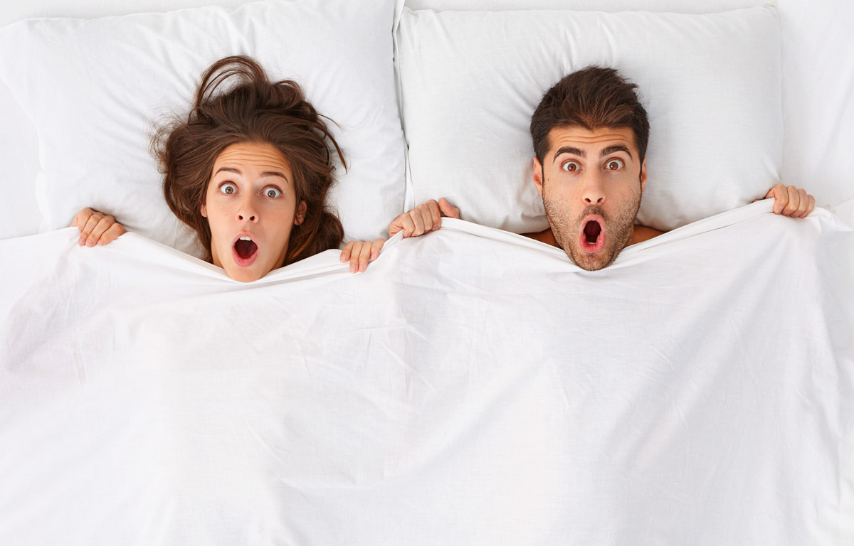 Zwei Personen liegen nebeneinander im Bett und schauen mit weit aufgerissenen Augen und geöffnetem Mund überrascht nach vorn, während sie die Bettdecke bis zum Kinn hochziehen.