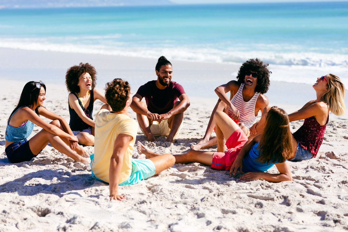 Gruppe junger Menschen sitzt lachend im Kreis am Strand vor dem Meer