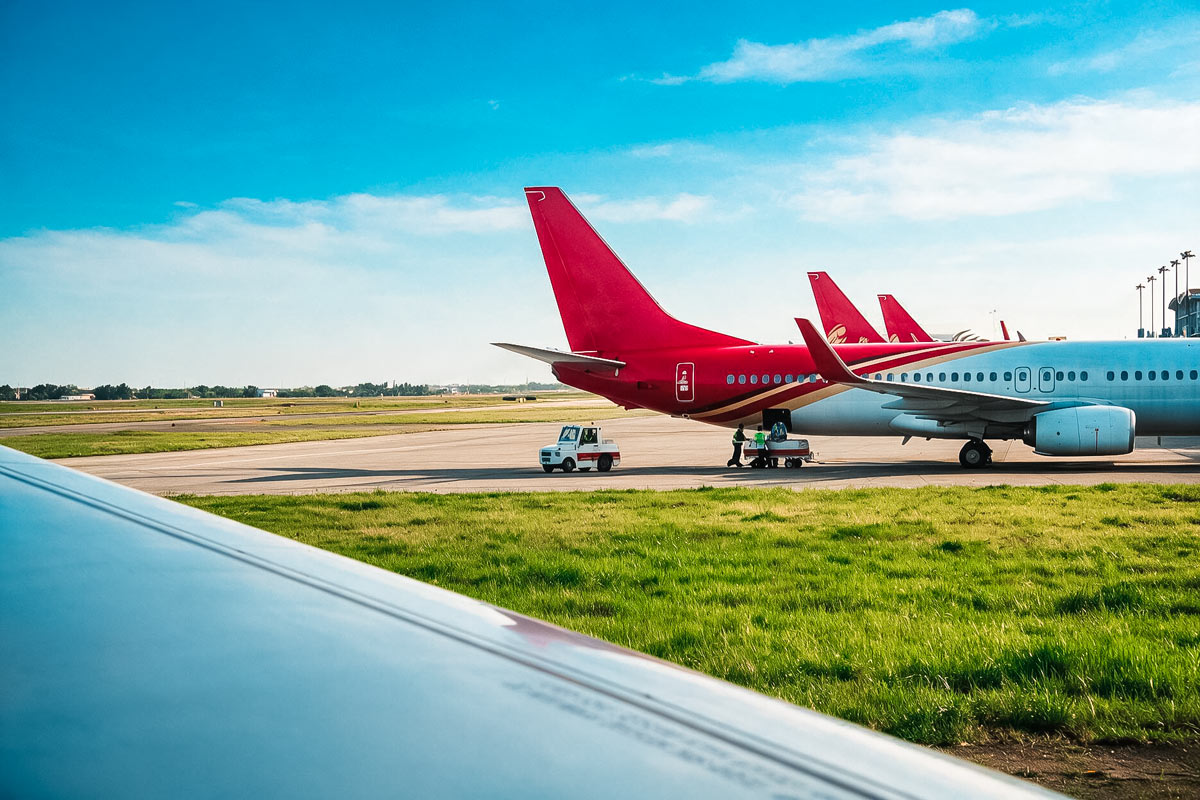 Ein Verkehrsflugzeug steht auf dem Rollfeld eines Flughafens. Im Vordergrund ist ein Teil einer Flugzeugtragfläche zu sehen. Auf dem Rollfeld befinden sich Fahrzeuge und Gepäckwagen. Das Wetter ist sommerlich.