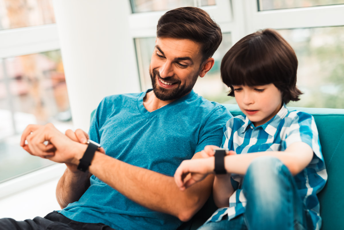 Mann und Junge sitzen nebeneinander auf einem Sofa und schauen lächelnd auf ihre Smartwatches am linken Handgelenk