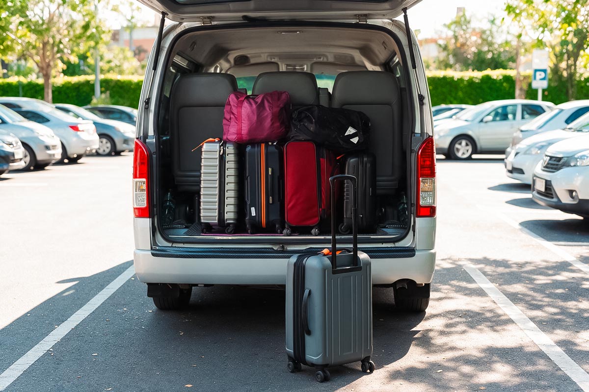 Offener Kofferraum eines Vans mit mehreren Koffern und Taschen, davor ein Trolley auf dem Parkplatz