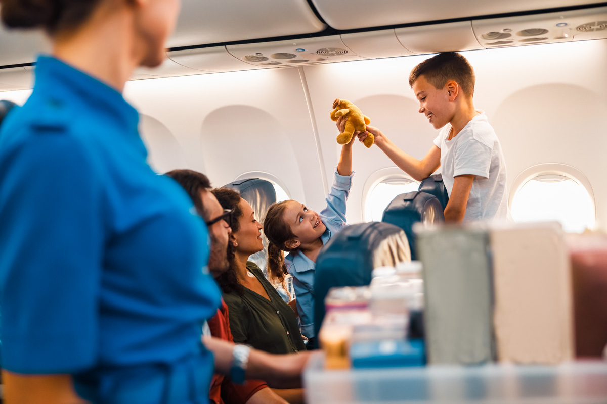 Kind überreicht im Flugzeug einem anderen Kind ein Stofftier, während Erwachsene zuschauen und eine Flugbegleiterin im Vordergrund steht