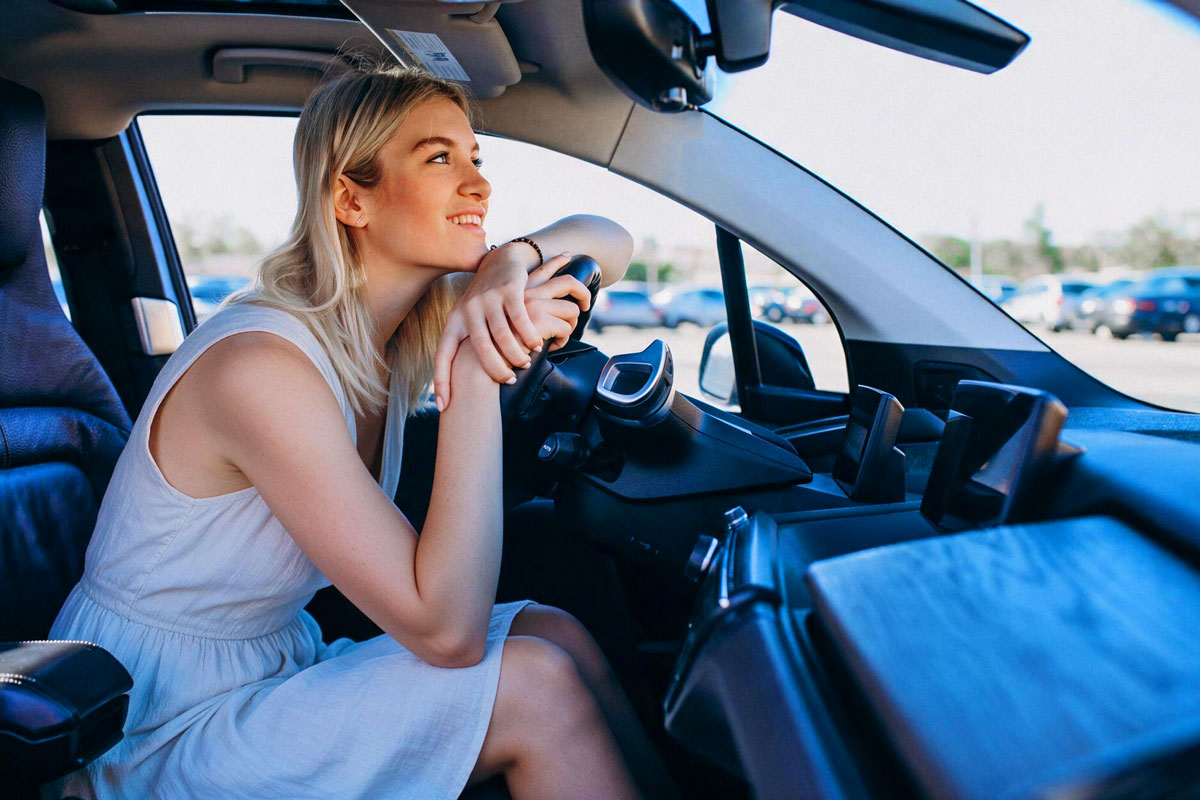 Junge Frau in einem Sommerkleid sitzt lächelnd am Steuer eines Autos auf einem Parkplatz
