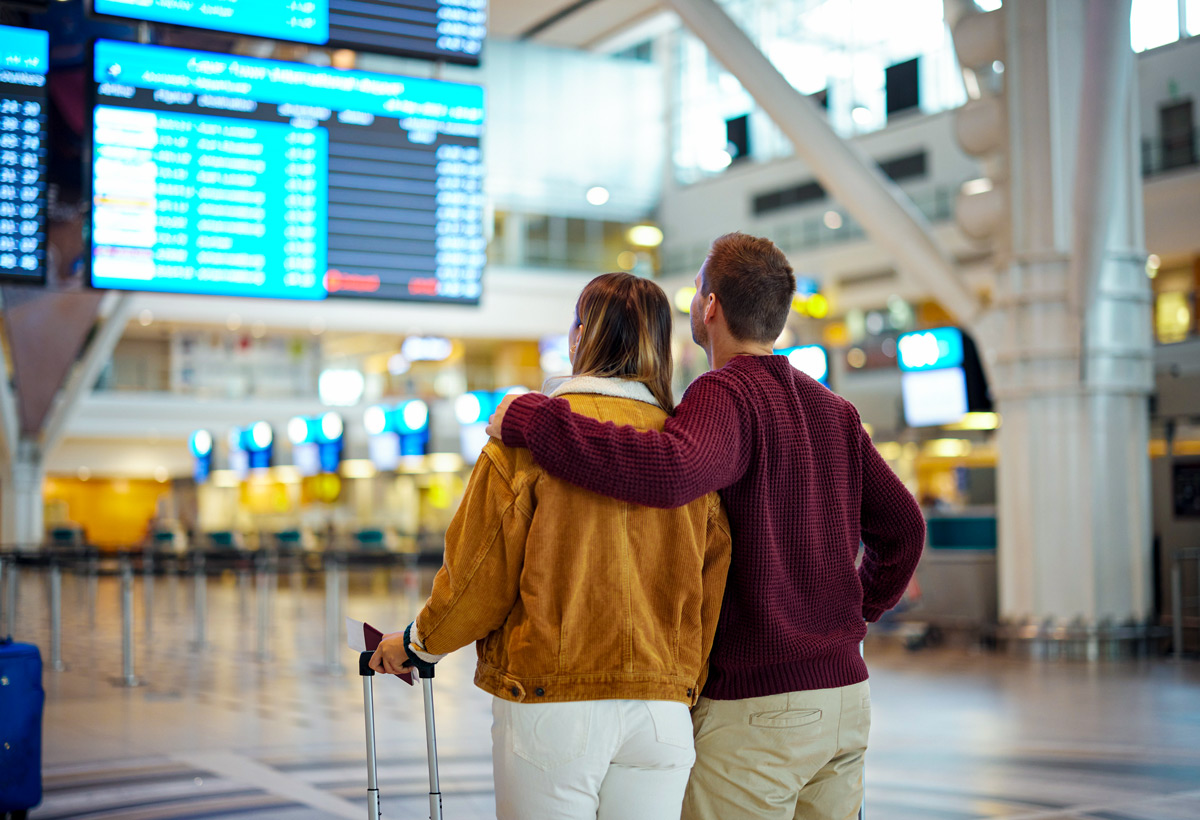 Ein Paar steht mit einem Rollkoffer in einem hellen Flughafen vor einer großen Anzeigetafel