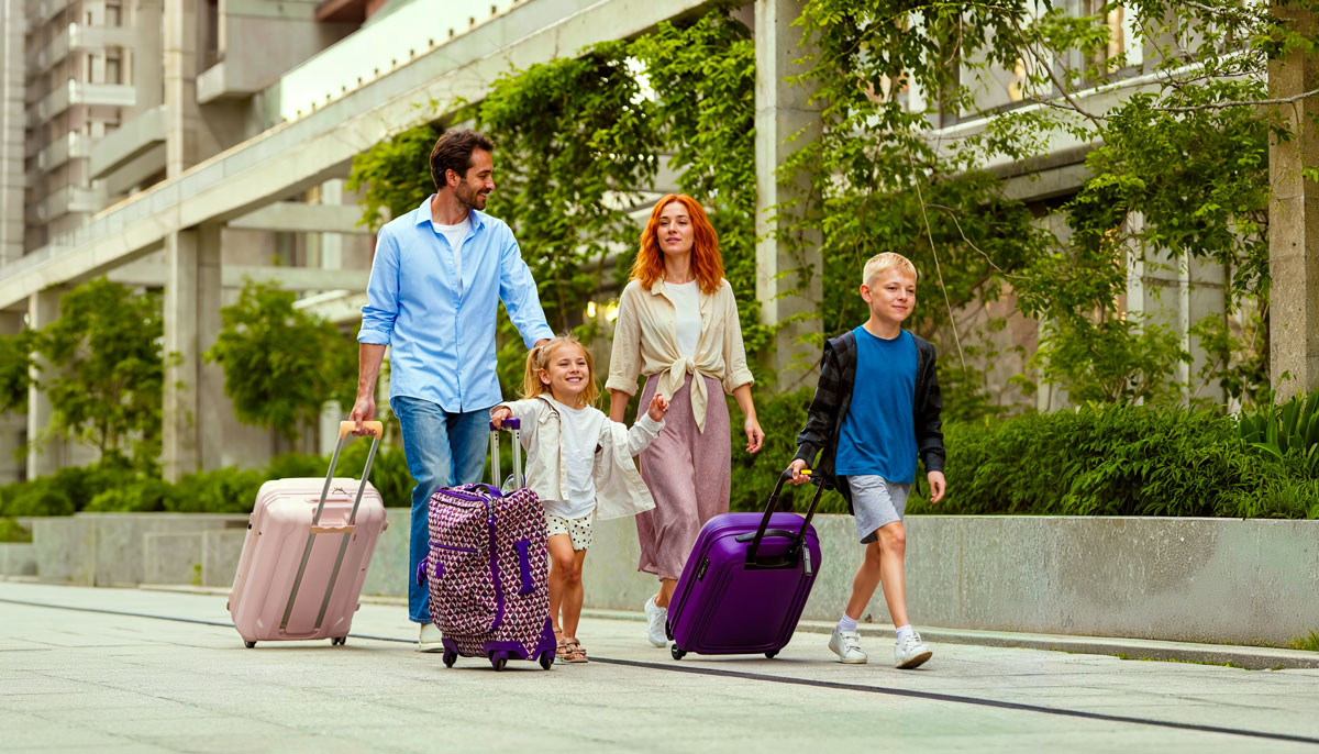 Eine Familie mit vier Personen geht nebeneinander auf einem Gehweg entlang. Alle ziehen Rollkoffer hinter sich her und lächeln glücklich. 
