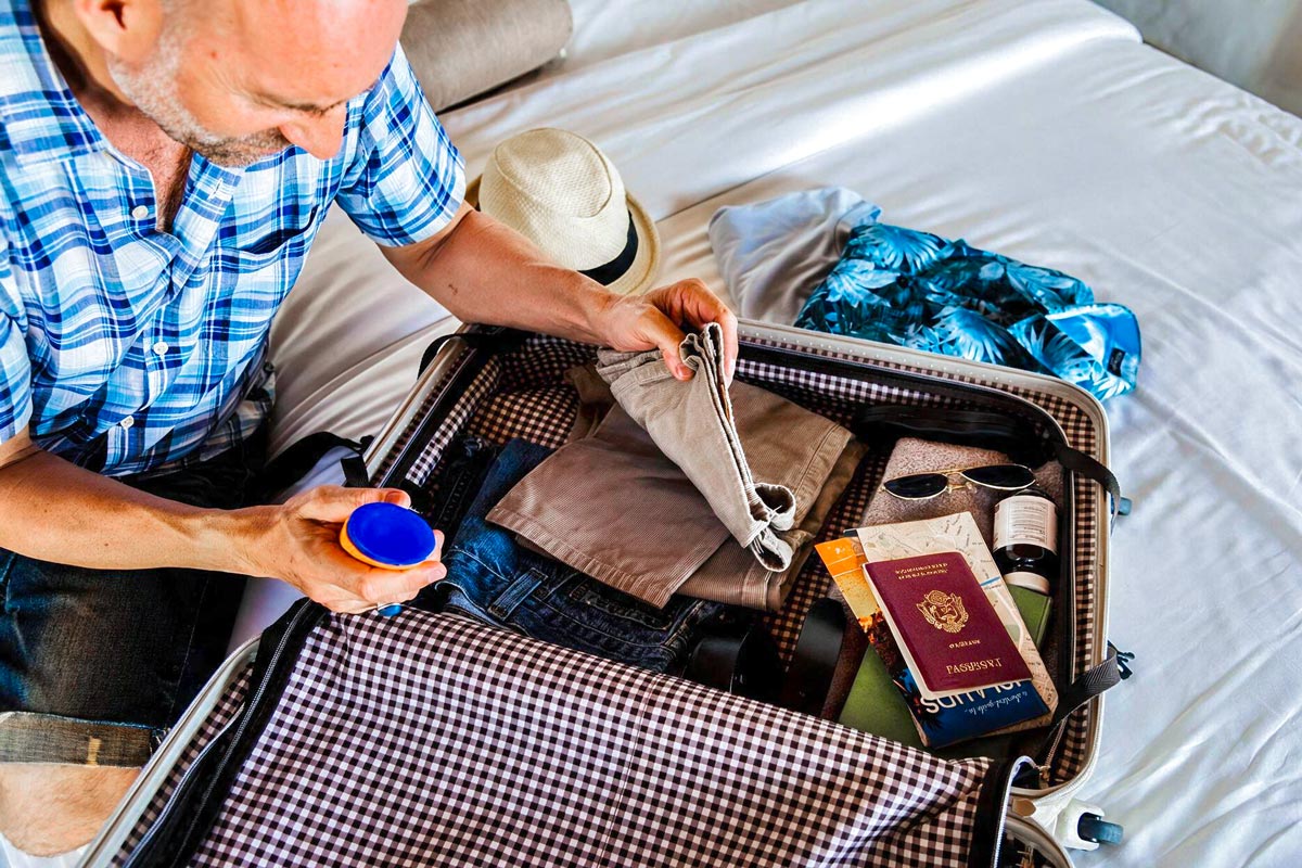 Ein Mann packt einen offenen Koffer auf einem Bett aus. Im Koffer liegen Kleidung, ein Reisepass, eine Sonnenbrille, ein Sonnenhut, ein Fläschchen Sonnencreme und weitere Reiseutensilien. 