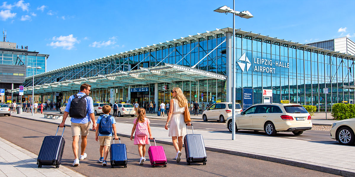 Zwei Erwachsene und zwei Kinder gehen mit Rollkoffern zum verglasten Terminal des Flughafens Leipzig/Halle.