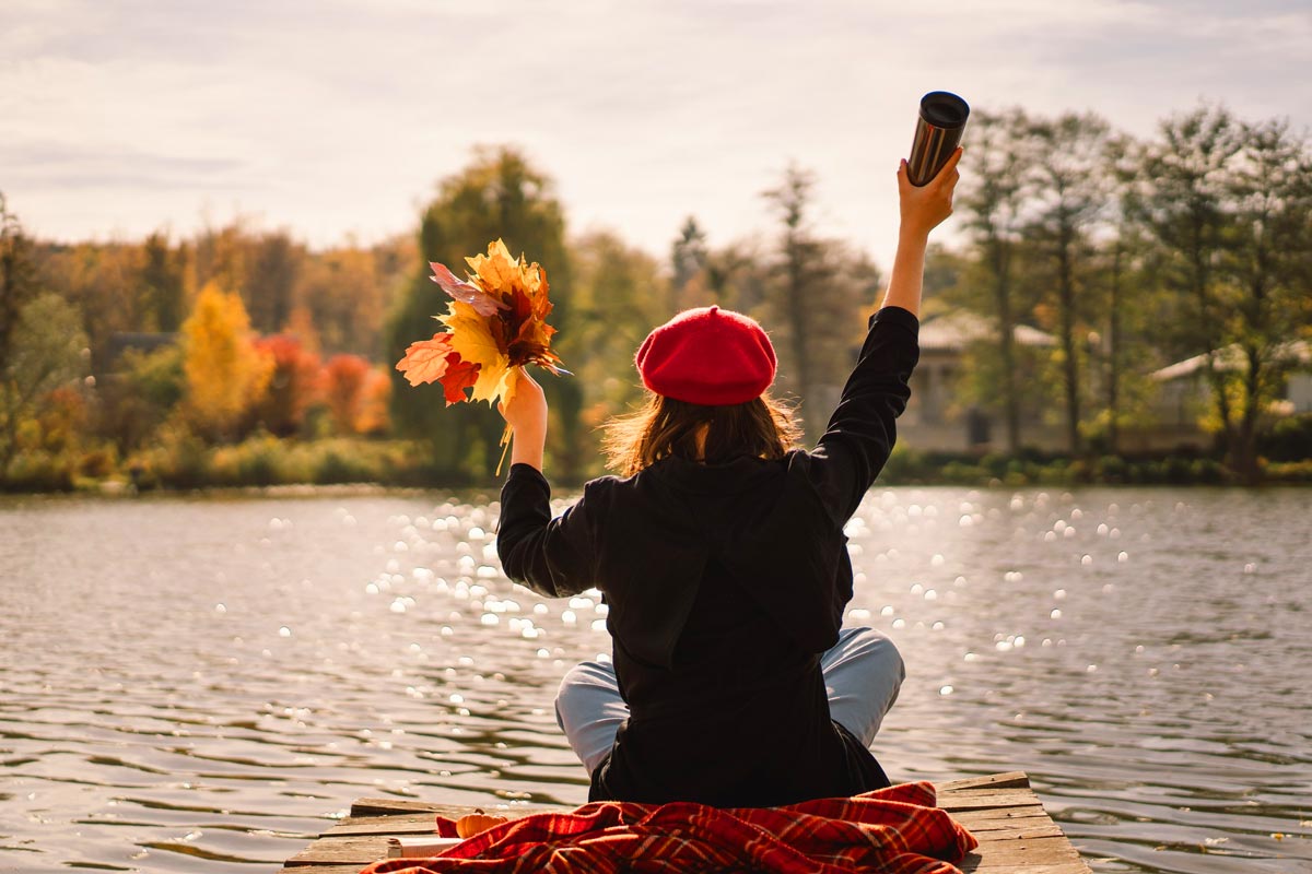 Person mit roter Baskenmütze sitzt auf einem Steg am See und hält einen Becher und einen Strauß bunter Herbstblätter in die Luft