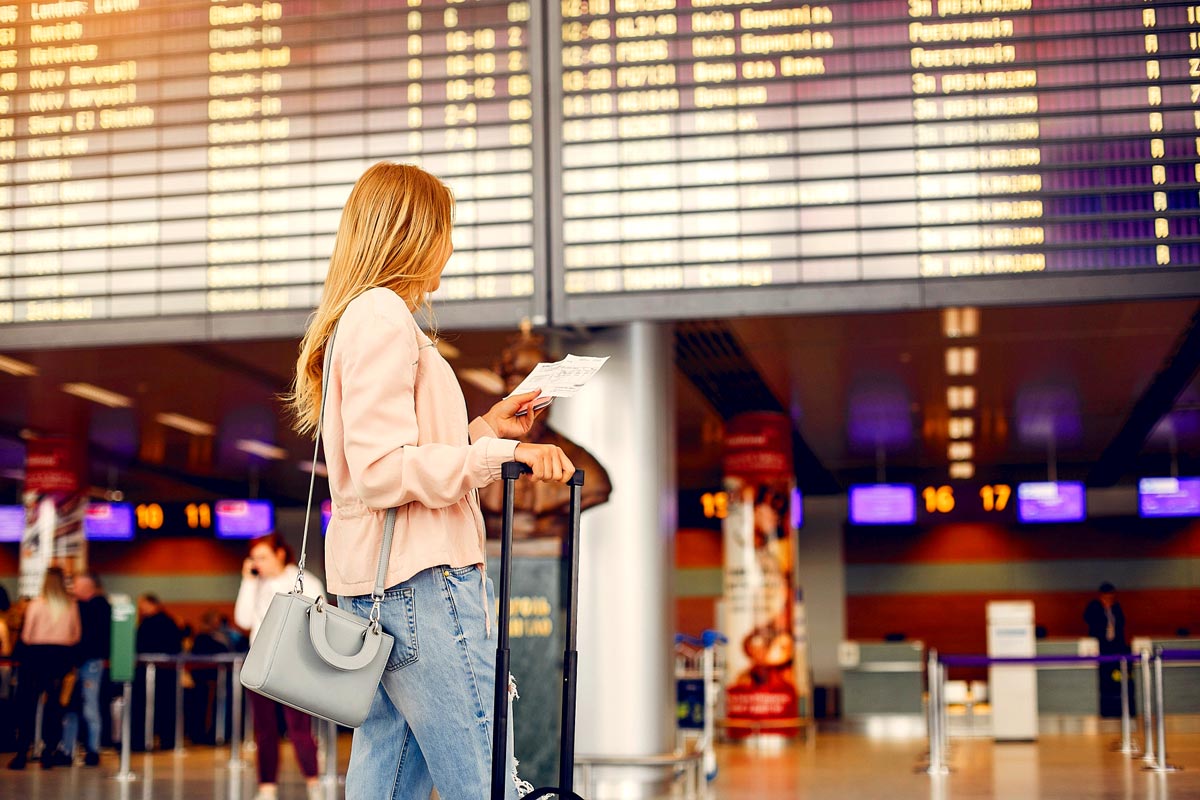 Frau mit Handgepäck und Tasche steht vor einer großen Anzeigetafel im Flughafen und hält Papiere in der Hand