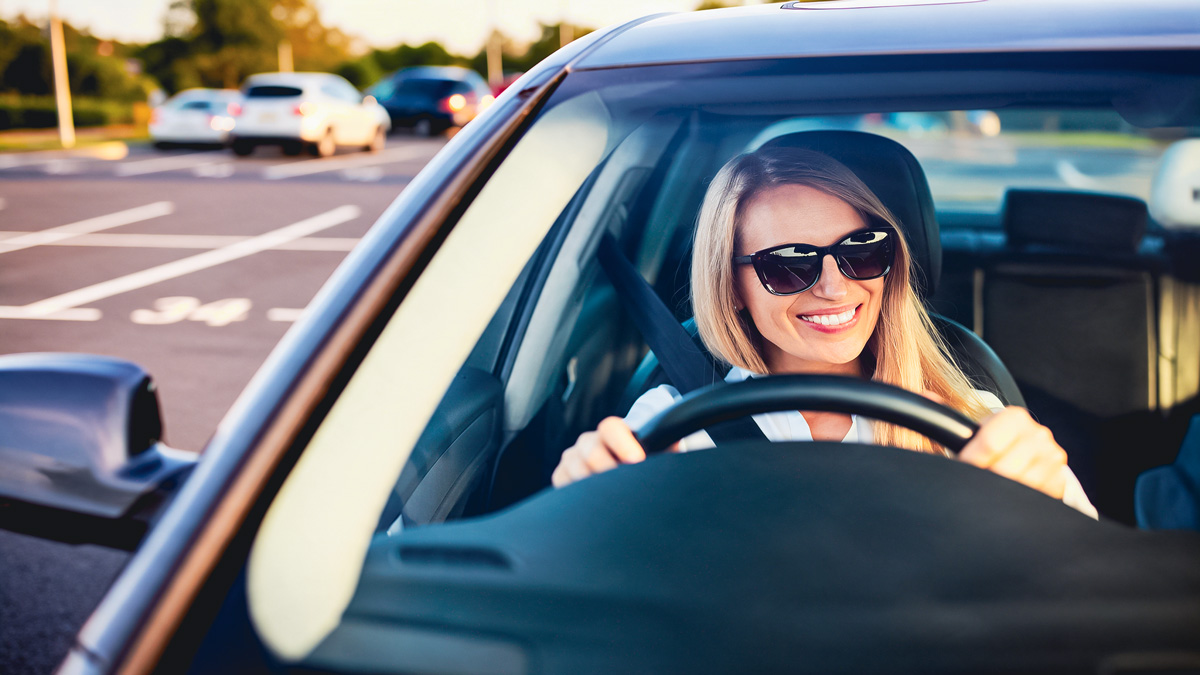 Frau mit Sonnenbrille sitzt lächelnd am Steuer eines Autos auf einem Parkplatz