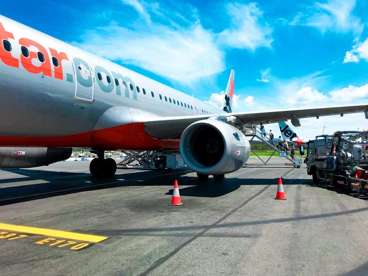 Flugzeug steht am Boden mit sichtbarem Triebwerk, Flügel und Bodenfahrzeugen auf dem Rollfeld, während Passagiere bei sommerlichem Wetter einsteigen.