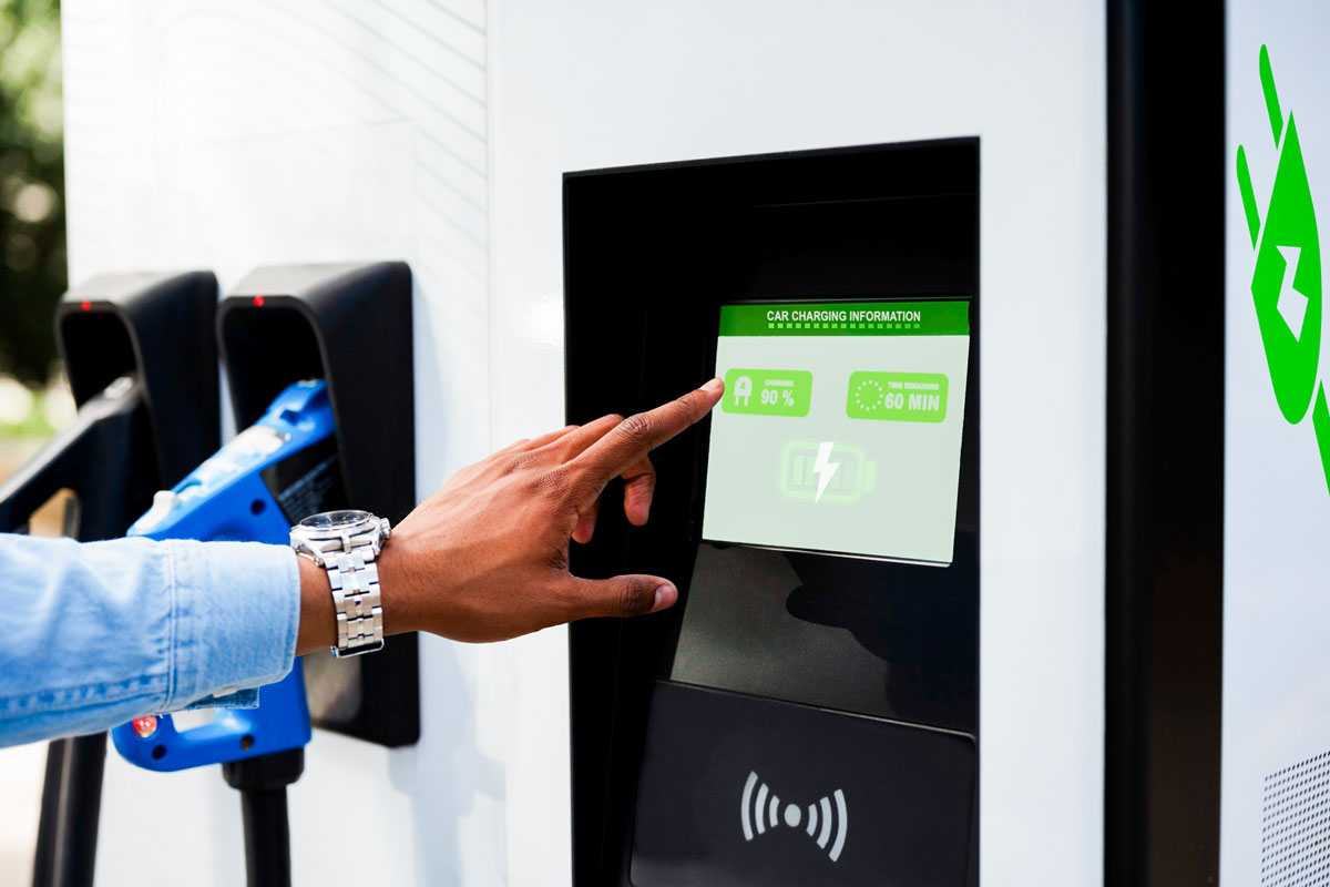 Hand tippt auf Display einer Ladestation für Elektrofahrzeuge mit Ladesteckern im Hintergrund