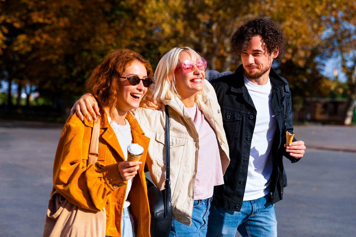 Drei junge Erwachsene gehen Arm in Arm im Freien und essen Eis bei sonnigem Herbstwetter