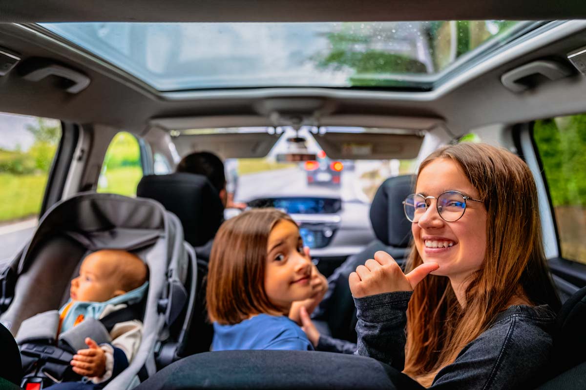 Drei Kinder sitzen auf der Rückbank im Auto, zwei schauen nach hinten und eines nach vorne, eines trägt eine Brille und lächelt. Der Vater sitzt am Lenkrad.