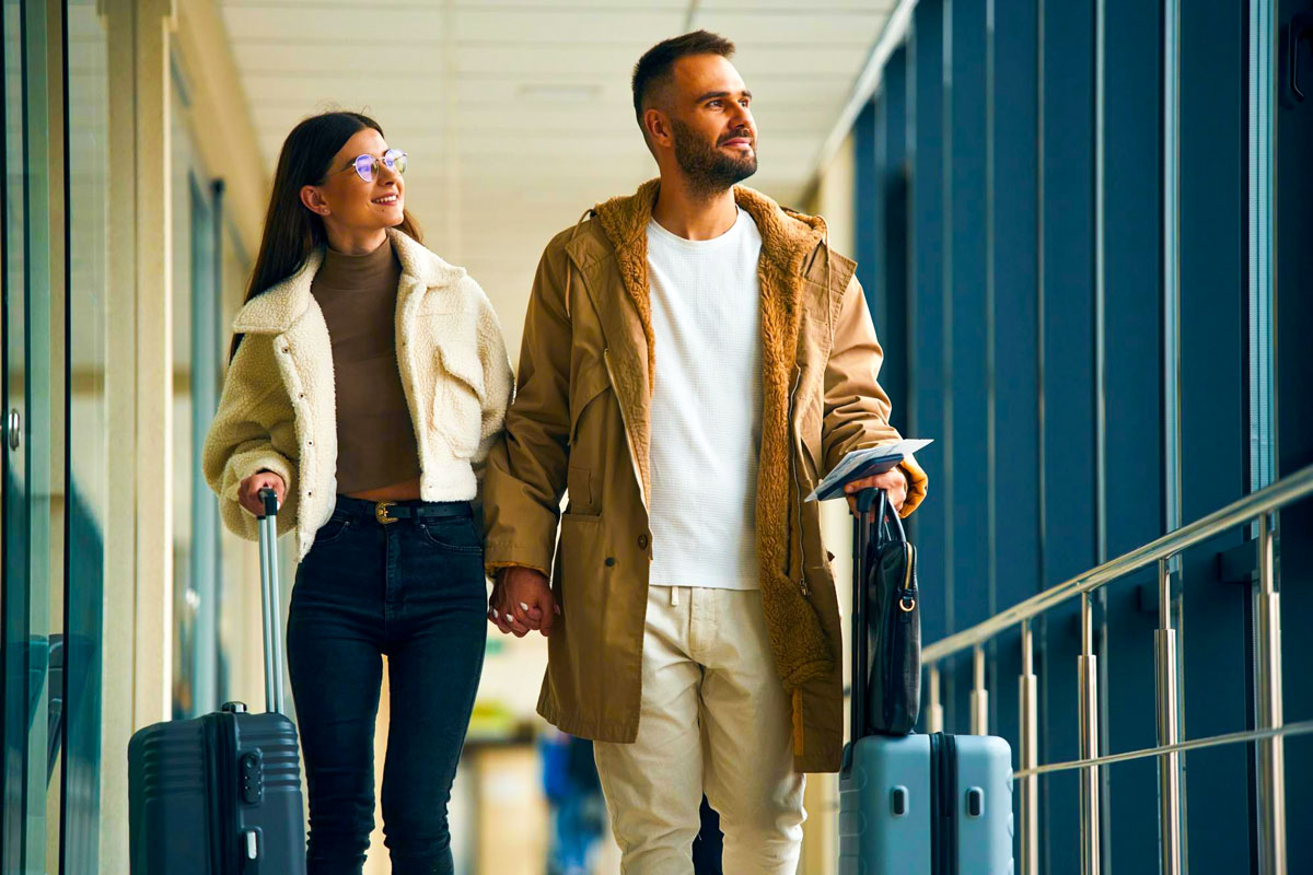 Paar mit Winterjacken und Rollkoffern geht Hand in Hand durch einen hellen Gang am Flughafen