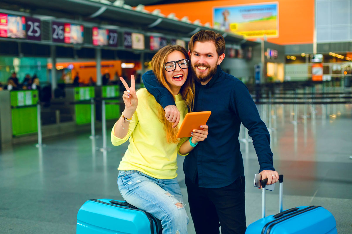 Junges Paar mit Koffern posiert lachend und zeigt Victory-Zeichen in einem Flughafen-Terminal