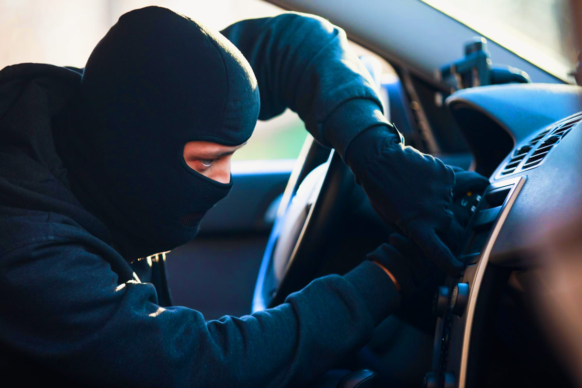 Person mit schwarzer Sturmhaube und Handschuhen sitzt im Auto und manipuliert das Armaturenbrett