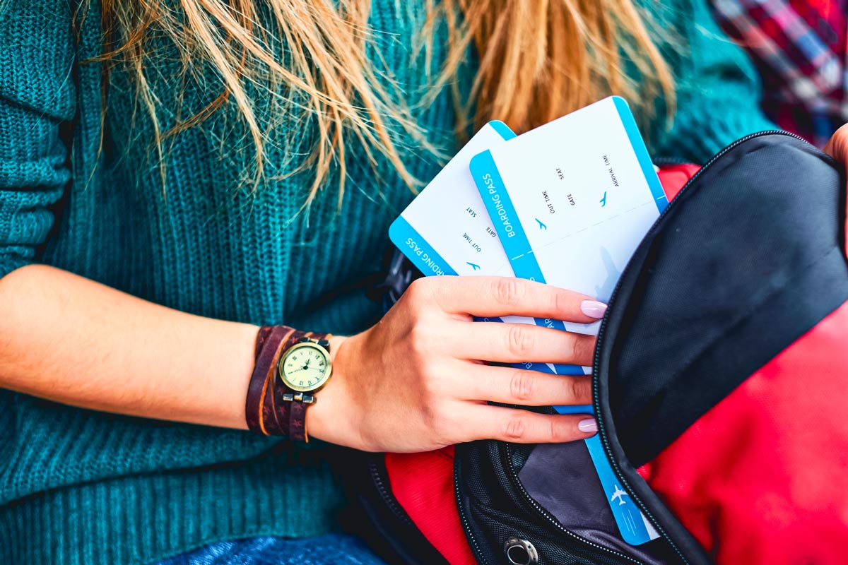 Hand mit Armbanduhr steckt zwei Flugtickets in einen Rucksack