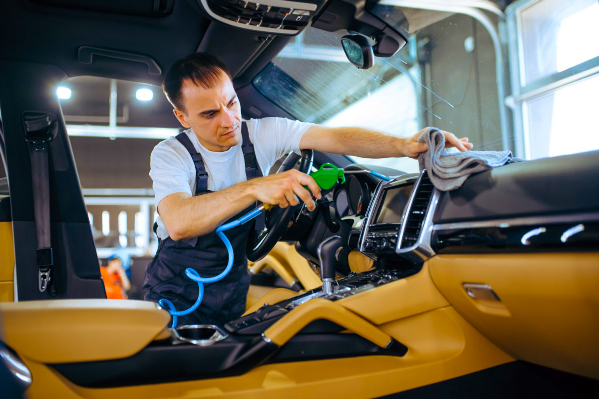 Mann in Arbeitskleidung reinigt mit Druckluftpistole und Tuch das Armaturenbrett eines Fahrzeugs mit modernem Innenraum